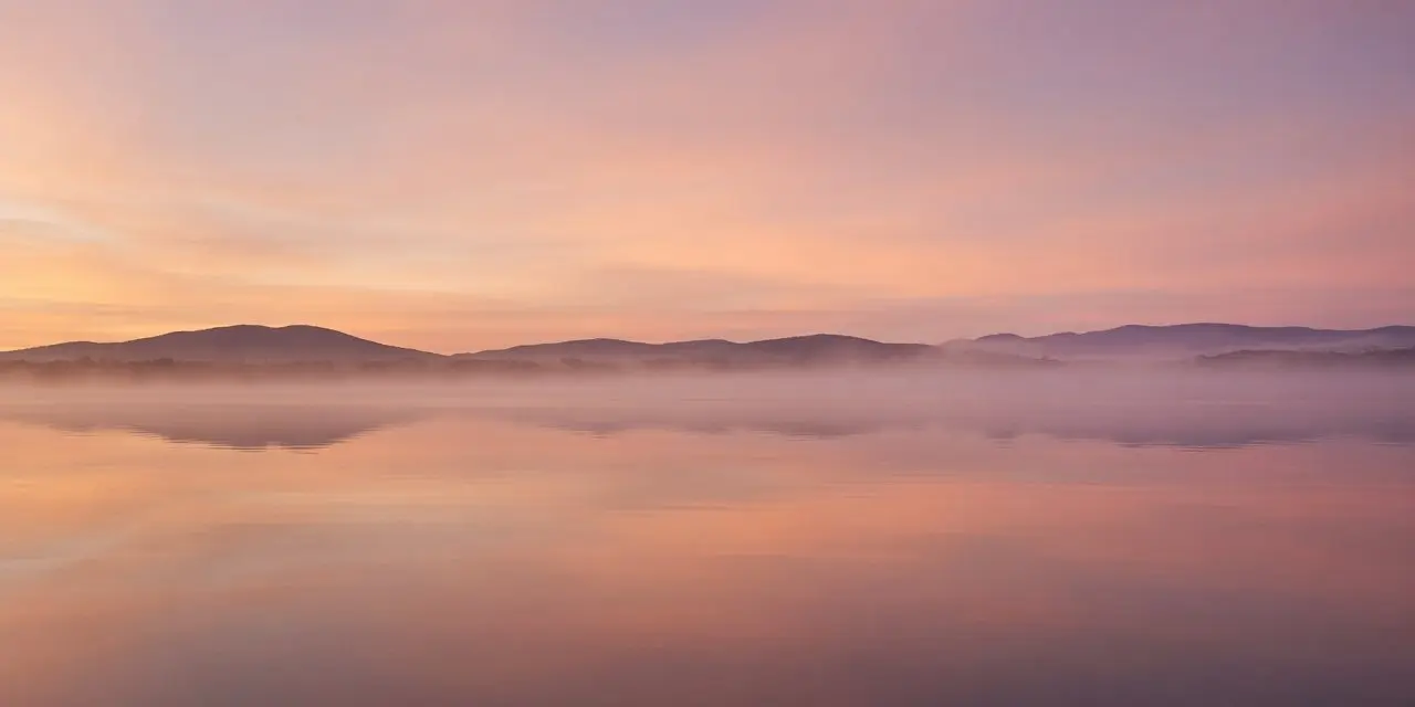 Calm, misty lake at dawn with soft peach and plum skies over distant hills